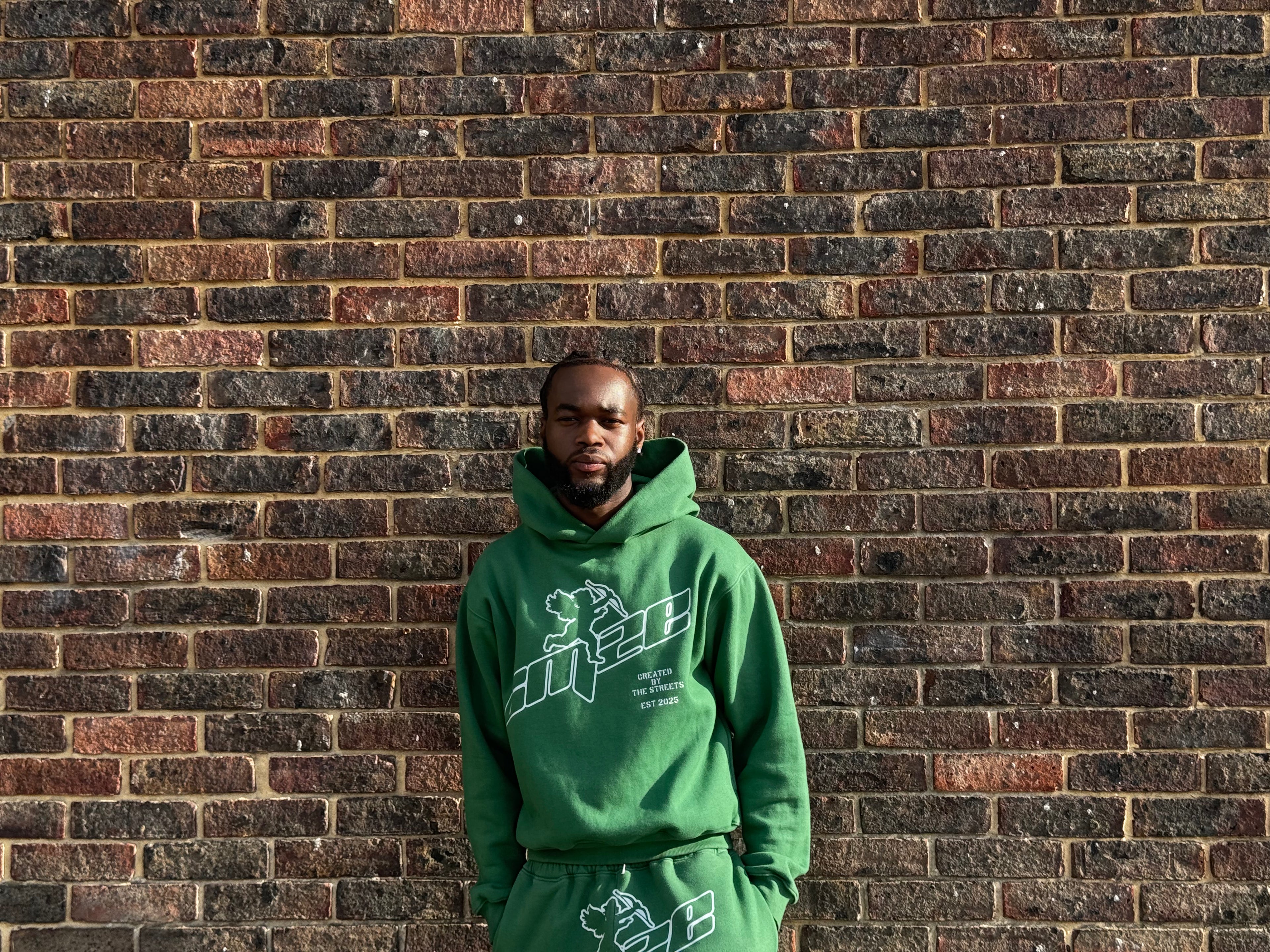 Young black man wearing a green tracksuit with a logo against a brick wall. The logo is CMZE