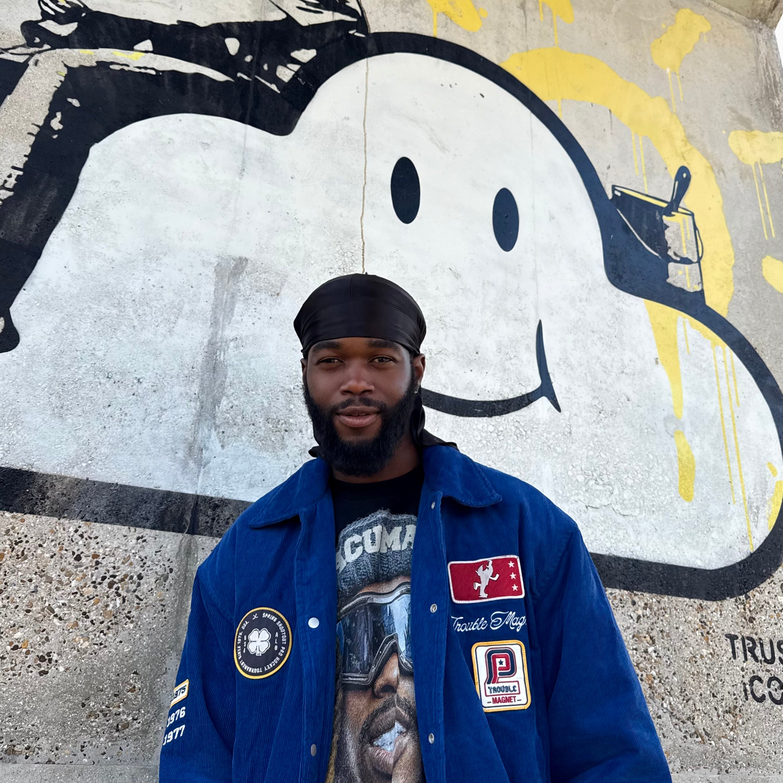 Man wearing a blue jacket with patches in front of a graffiti wall.